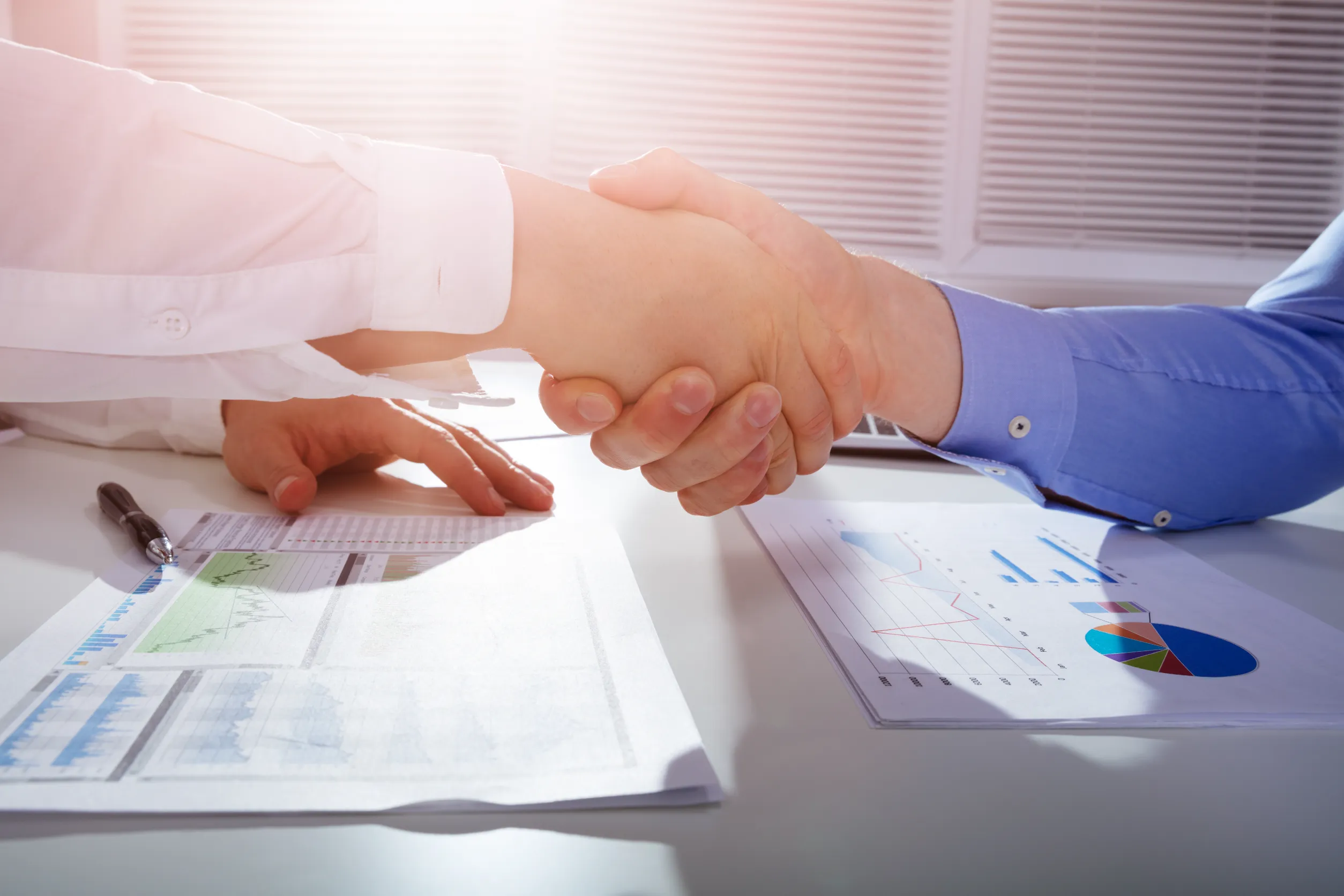 Close-up Of Two Businessmen Shaking Hands Over Financial Graph Report On White Desk In Office
